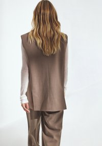 Braun ärmellose Weste mit geradem Schnitt, kombiniert mit lockeren, hellbraunen Hosen. Strukturiertes Material; sanfte, neutrale Farbpalette.