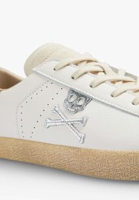 White leather sneaker with beige suede accents, featuring a silver skull and crossbones embroidery, perforated design, and textured rubber sole.