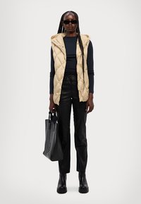 Beige quiltet ærmeløs vest, marineblå langærmet top, sorte tætsiddende bukser, sorte ankelstøvler og en sort tote bag. Glat tekstur overalt.