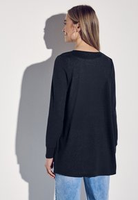 Svart långärmad tunika med rund halsringning, sidoöppningar och en mjuk textur, kombinerad med ljusblå denim. Minimalistiska designelement.
