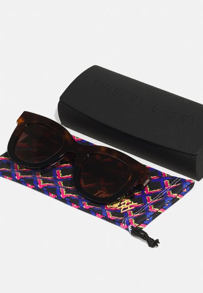 Des lunettes de soleil en écaille brune sur une pochette à motifs bleus et roses, accompagnées d'un étui rigide noir embossé avec "THIERRY LASRY."