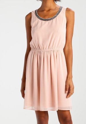 Robe rose clair sans manches avec taille froncée et détail de décolleté orné de perles, portée par une personne aux bras et aux jambes supérieures visibles.