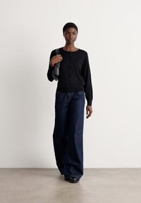 Svart cardigan med knappelukking, teksturert stoff, kombinert med løstsittende mørk denim bukser og svarte sko, minimalistisk design.
