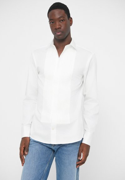 Bela srajca z dolgimi rokavi iz gladkega materiala, z nabranim sprednjim delom in koničastim ovratnikom, v kombinaciji z modrimi kavbojkami.