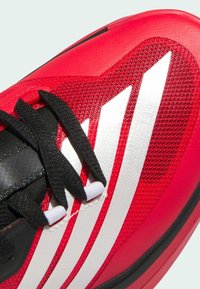 Chaussure de sport rouge avec des lacets noirs et trois bandes blanches, dotée d’une tige en mesh texturé et en matériau synthétique.