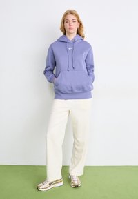 Fato de capuz de algodão roxo com um bolso frontal e cordão, combinado com calças largas brancas e ténis com detalhes prateados sobre um chão verde.