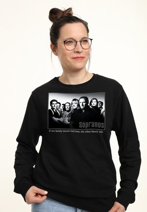 Kobieta w okularach nosząca czarną bluzę z obsadą serialu The Sopranos oraz cytatem "If one family doesn't kill him, the other family will."