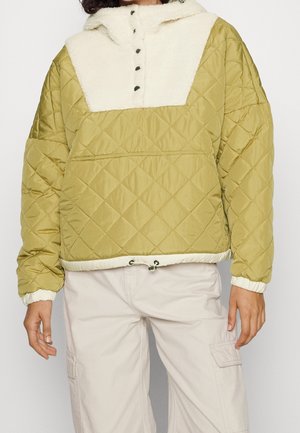 Persoon draagt een doorgestikte olijfgroene trui met crèmekleurige fleece kraag en knopen aan de voorkant, gecombineerd met lichtbeige cargobroek.