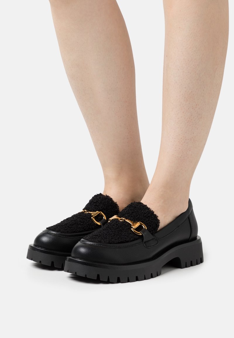 Jambes portant des mocassins noirs épais avec un tissu supérieur texturé et un détail en métal doré en forme de mors de cheval sur un fond blanc.