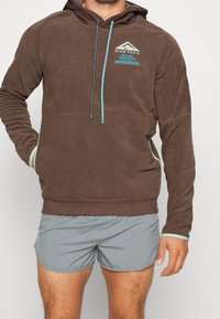 Felpa in pile marrone con cappuccio regolabile, dotata di accenti turchesi e tasca frontale. Indossata con pantaloni corti grigi.