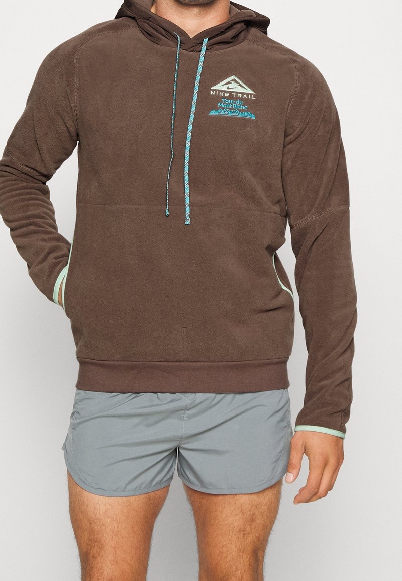 Felpa in pile marrone con cappuccio regolabile, dotata di accenti turchesi e tasca frontale. Indossata con pantaloni corti grigi.