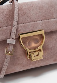 Vaaleanpunainen mokkanahkainen läppälaukku, jossa on kiiltävä kultainen lukitusmekanismi ja läpinäkyvä yksityiskohta. Sisältää ohuen hihnan ja tekstuuripinnan.