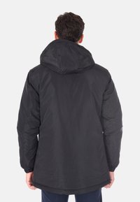 Uomo che indossa un giubbotto invernale nero con cappuccio, visto di spalle, in piedi su uno sfondo bianco semplice.