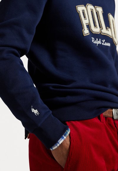 Navy Sweatshirt mit "POLO" in cremiger Farbe gestickt. Weißes Polo-Logo am Ärmel, kombiniert mit roten Cordhosen und einem karierten Hemdumschlag.
