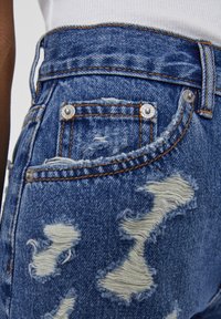 Primer plano de unos vaqueros de mezclilla azul desgastados con agujeros deshilachados, costuras visibles, remaches de metal y un bolsillo lateral en una persona que lleva una camiseta blanca.