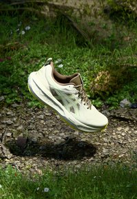 Leichter beiger Trailrunning-Schuh mit strukturiertem Obermaterial, Mesh-Einsätzen, grünen Akzenten und einer gepolsterten Sohle, die über dem Boden schwebt.