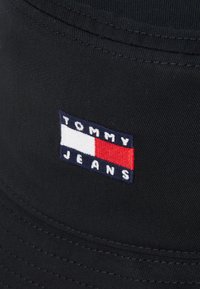 Juodas kibirėlio skrybėlė, pagaminta iš tvirto audinio, su siuvinėtu logotipo pleistru, kuriame užrašyta „TOMMY“ balta spalva ir „JEANS“ jūros mėlyna spalva, akcentuota raudona ir balta juosta.