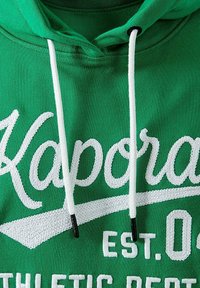 Hoodie vert avec texte brodé blanc "Kaporal" et "EST. 04," doté de cordons blancs avec embouts en plastique noir. Texture douce.