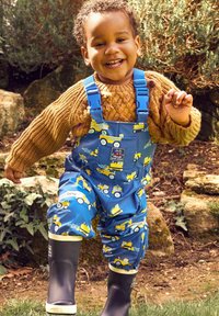 Modré nepremokavé nohavice s motívmi žltých stavebných vozidiel, nastaviteľné modré popruhy a pod nimi textúrovaný hnedý pletený sveter.