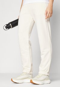 Pantalons de jogging blancs avec des poignets élastiques, poches latérales et une coupe décontractée, associés à des baskets de couleur claire et une bouteille d'eau noire.