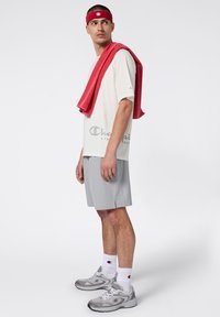 Męski strój sportowy składający się z jasnoszarej koszulki z krótkim rękawem z logo, dopasowanych szarych szortów oraz czerwonego ręcznika zarzuconego na ramię.