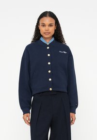 Námornícky skrátený sveter s okrúhlym golierom, zlatými gombíkmi, bočnými vreckami a vyšitým logom. Vyrobený z jemnej látky s hladkou textúrou.