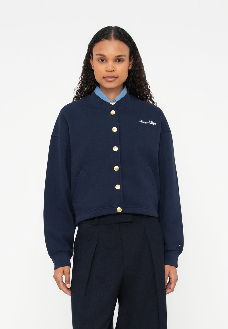 Námornícky skrátený sveter s okrúhlym golierom, zlatými gombíkmi, bočnými vreckami a vyšitým logom. Vyrobený z jemnej látky s hladkou textúrou.