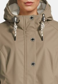 Femme portant une veste à capuche beige avec doublure en polaire blanche, boutons-pression à l'avant, et cordons blancs avec embouts noirs.