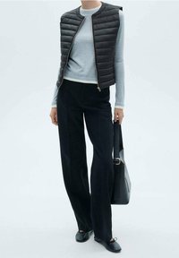 Czarna pikowana kamizelka noszona na jasnoszarej koszuli z długim rękawem, zestawiona z czarnymi dopasowanymi spodniami i czarnymi płaskimi butami. Do tego duża czarna torba na ramię.