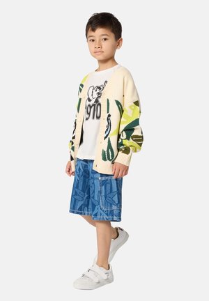 Gebreid vest met groene en gele bladerenpatronen over een wit T-shirt met een olifantafbeelding, gecombineerd met blauwe korte broek met patronen en witte schoenen.