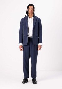 Traje navy con un blazer ajustado y pantalones a juego; camisa blanca de botones por debajo; textura suave y diseño clásico; zapatos negros.