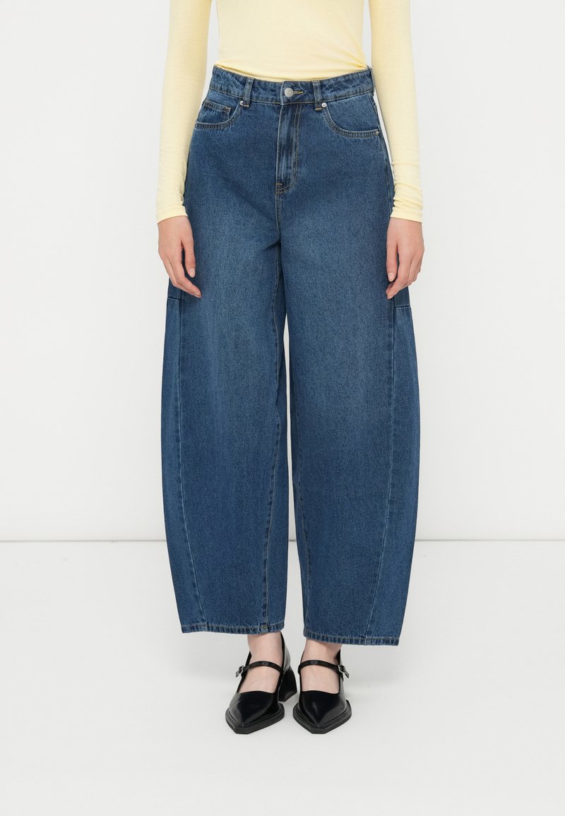 Jeans de mezclilla de corte ancho y color azul claro con cintura alta, cierre de botón y cremallera, y bolsillos frontales, combinados con zapatos negros planos.