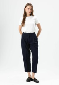 Witte korte mouwen blouse gecombineerd met hooggetailleerde, marineblauwe broek met plooien. De broek heeft een omgeslagen zoom. Het model draagt zwarte loafers.