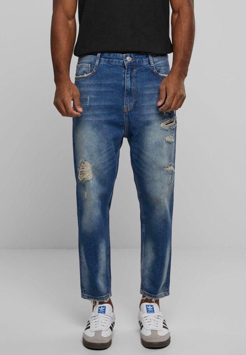 Jeans de mezclilla azul con un diseño desgastado, que presenta detalles rasgados, un ligero desvanecimiento y un corte recto. Combinados con zapatillas blancas.