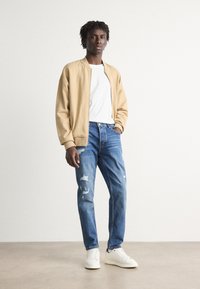 Beige bomber jacketin päällä valkoinen t-paita, yhdistettyinä kuluneisiin sinisiin farkkuihin ja valkoisiin lenkkareihin. Sileä kangas, rento leikkaus, moderni ulkonäkö.