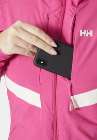 Světlá růžová bunda s bílými akcenty, zipovou kapsou a černým telefonem, který se zasouvá. Má hladkou tkaninu a kontrastní zip.