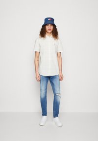 Camisa de cuadros de manga corta en beige claro, gorra tipo bucket azul con logo, jeans azules desgastados y zapatillas blancas, de pie contra un fondo plano.