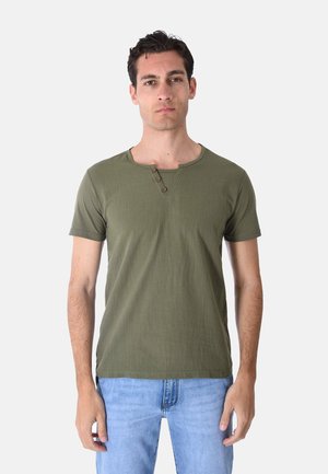 Giovane uomo con capelli corti e scuri che indossa una maglietta henley verde oliva a maniche corte e jeans azzurri chiari, in piedi davanti a uno sfondo semplice.