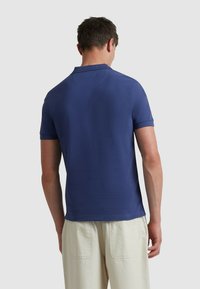 Polo shirt bleu marine à manches courtes avec col, texture lisse et coupe droite, associé à un pantalon de couleur claire dans un cadre décontracté.