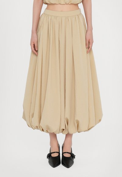 Beige, mid-calf skirt with a gathered design and voluminous shape. Features an elastic waistband and is paired with black, pointed-toe shoes.