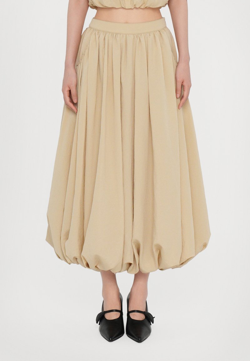 Beige, knielanger Rock mit gerafftem Design und voluminöser Form. Verfügt über einen elastischen Bund und wird mit schwarzen, spitz zulaufenden Schuhen kombiniert.