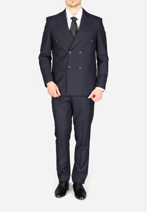 Homme portant un costume croisé bleu marine avec cravate noire, chemise blanche, pantalon assorti et chaussures de ville noires, debout sur un fond blanc.