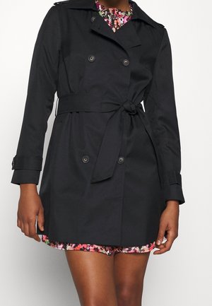 Kobieta ubrana w czarny, dwurzędowy trencz z paskiem zawiązanym w talii na kolorowej, kwiecistej sukience.