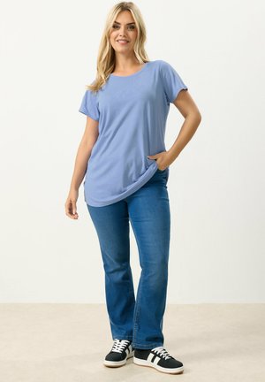 Mujer sonriendo, con camiseta de manga corta azul claro, vaqueros azules y zapatillas negras con rayas blancas, de pie frente a un fondo liso.