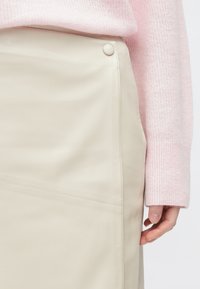 Béžová sukně z umělé kůže s elegantním designem, zdobená viditelným švem a knoflíkovým akcentem, doplněná měkkým růžovým pleteným svetrem.