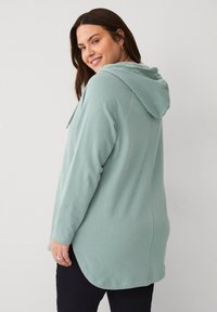 Sudadera verde claro con cordón, mangas largas y un bajo trasero ligeramente más largo, fabricada con un tejido suave y texturizado.