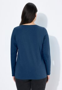 Kobieta z długimi ciemnymi włosami, ubrana w prostą granatową bluzkę z długim rękawem i czarne spodnie, stojąca przodem do białej ściany z widocznym cieniem.