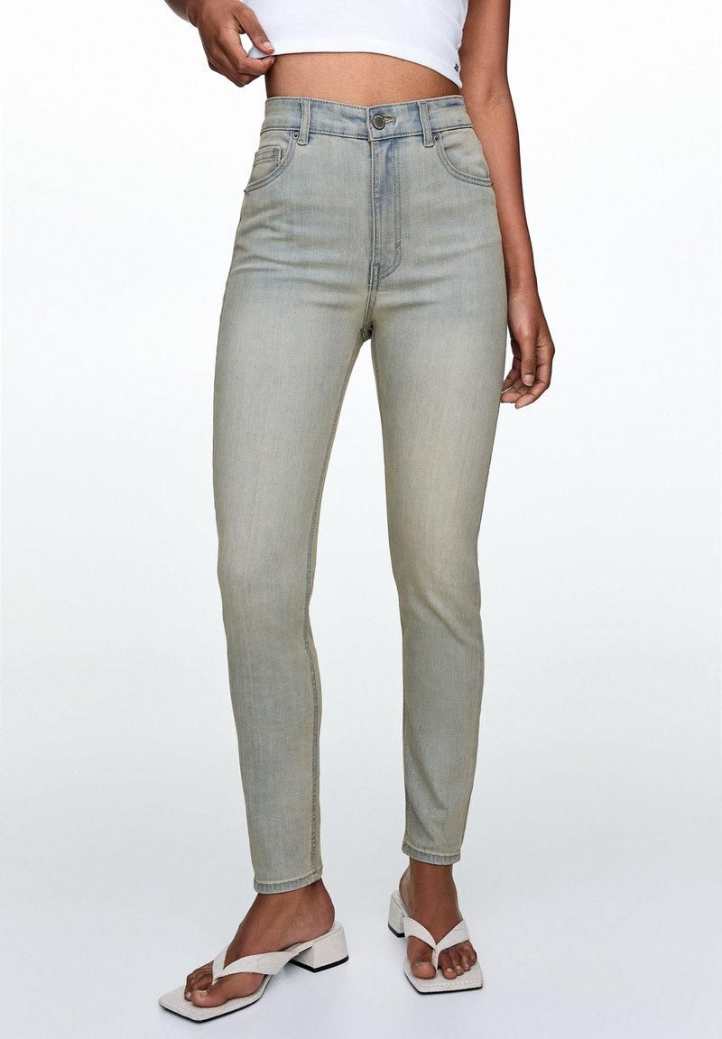 Jeans cintrés taille haute de couleur bleu clair, coupe slim, cinq poches, fermeture par bouton métallique, avec un léger effet délavé. Associés à des sandales blanches à talons.