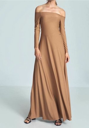 Vrouw in een lange, beige off-shoulder jurk met lange mouwen en zwarte open-toe hakken, staand tegen een effen lichte achtergrond.