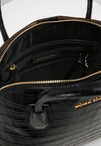 Czarna torebka skórzana o fakturze krokodyla z złotym suwakiem i logo Michael Kors, pokazująca wnętrze z zasuwaną kieszenią i czarną podszewką.